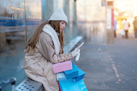 Eine lächelnde junge Frau mit bunten Einkaufstüten, die sich in einer urbanen Winterstadt mit ihren Handys im Freien vergnügt. Menschen, Kommunikation, Shopping und Lifestylekonzept