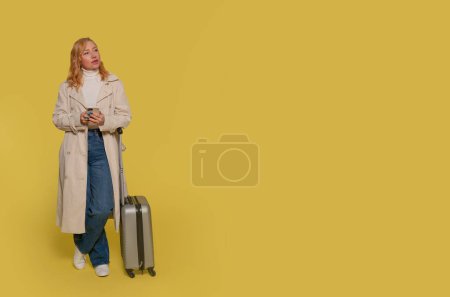 Eine Frau in Jeans, weißem Hemd und Trenchcoat trägt einen Koffer und ein Mobiltelefon auf gelbem Hintergrund. Glückliche Menschen im Urlaub, Urlaub