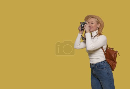 Eine Frau in Jeans, weißem Hemd und Hut trägt einen Koffer und fotografiert mit einer Vintage-Kamera auf gelbem Hintergrund. Glückliche Menschen im Urlaub, Urlaub
