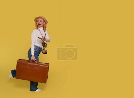 Eine Frau in Jeans, weißem Hemd und Hut trägt einen Koffer und fotografiert mit einer Vintage-Kamera auf gelbem Hintergrund. Glückliche Menschen im Urlaub, Urlaub