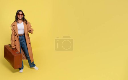 una mujer en jeans, una camiseta blanca y un abrigo marrón que lleva una maleta y se toma una selfie por teléfono sobre un fondo amarillo. Gente feliz yendo de vacaciones, vacaciones