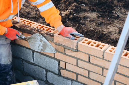 Industrial bricklayer laying bricks on cement mix on construction site. Fighting housing crisis by building more affordable houses concept
