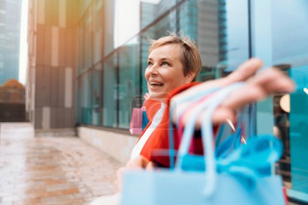 glückliche Frau in roter Jacke mit bunten Taschen, die Spaß beim Einkaufen in der Großstadt hat. Konsum, Verkauf, Einkauf, Einkaufen, Lifestylekonzept