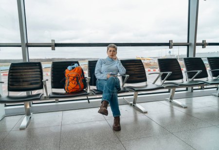 Reisende, die ein Telefon benutzen, auf einem Flughafen sitzen, auf einen Flug warten, einen Flughafen betreten und einen Rucksack bei sich tragen. Reisekonzept.