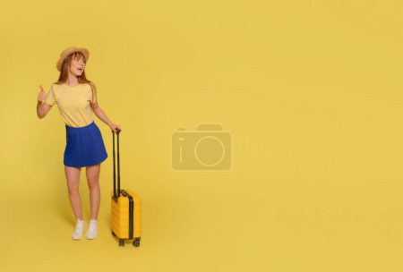 eine fröhliche junge Frau in blauem Rock, gelbem T-Shirt und Hut, die einen Koffer trägt und auf gelbem Grund den Daumen nach oben streckt. Glückliche Menschen im Urlaub, Urlaub