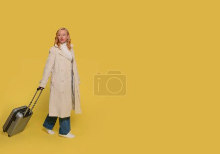 una mujer en jeans, una camisa blanca y una gabardina que lleva una maleta y usa un teléfono móvil sobre un fondo amarillo. Gente feliz yendo de vacaciones, vacaciones