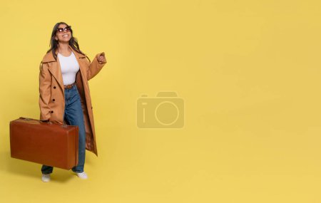 Eine Frau in Jeans, weißem T-Shirt und braunem Mantel trägt einen Koffer und macht ein Selfie mit dem Handy auf gelbem Hintergrund. Glückliche Menschen im Urlaub, Urlaub