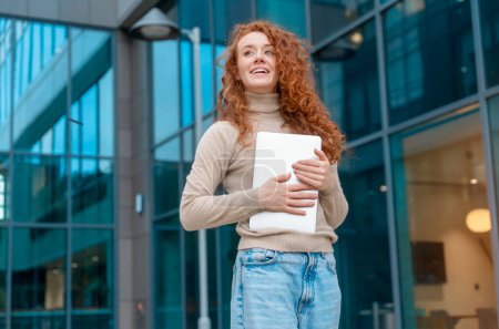 Portrait of beautiful happy young woman graduate with curly red hair holding laptop and excited  about news regarding her job application