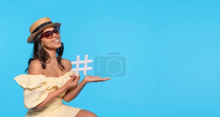 Mujer feliz en vestido amarillo y sombrero sosteniendo hashtag signo blanco en su mano sobre fondo azul. medios sociales, blogs y temas virales en internet felices vacaciones, concepto de vacaciones