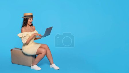 Mujer india en vestido amarillo sentada en la maleta usando portátil sobre fondo azul. Viajes disfrutar concepto
