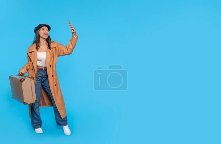 Eine Frau in Jeans, weißem T-Shirt und braunem Mantel trägt einen Koffer und macht ein Selfie per Telefon auf blauem Hintergrund. Glückliche Menschen im Urlaub, Urlaub