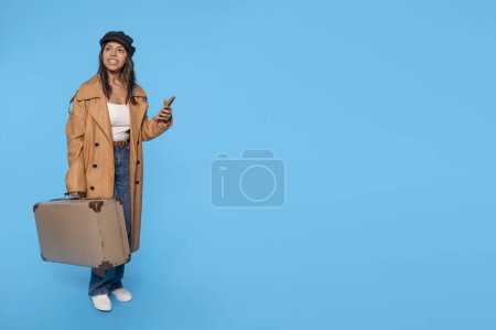 a woman in jeans, a white T-shirt, and a brown coat carrying a suitcase and using a mobile phone on a blue background. Happy people going on holiday, vacation