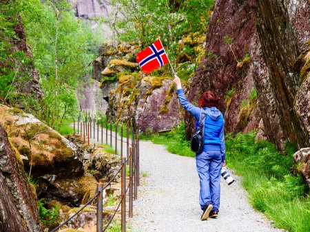 Mujer turista con bandera noruega y cámara caminando por el sendero de las montañas en Allmannajuvet área Sauda, Noruega. Atracción a lo largo de la ruta turística nacional Ryfylke.