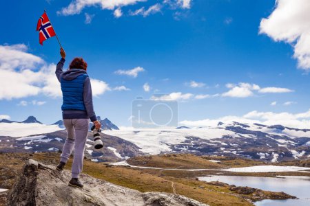 Tourist woman enjoy mountains landscape, holding norwegian flag and photo camera. National tourist scenic route 55 Sognefjellet, Norway