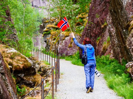 Tourist woman with norwegian flag and camera walking trough mountains path in Allmannajuvet area Sauda, Norway. Attraction along national tourist route Ryfylke.