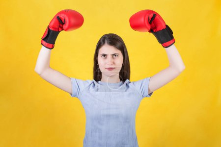 Photo for Confident young woman with red boxing gloves celebrating success, flexing arm muscles with hands raised up. Portrait of strong and determined girl isolated on yellow background. Self defence concept - Royalty Free Image