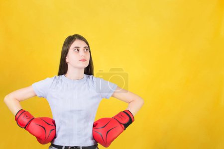 Photo for Confident young woman with red boxing gloves stands with hands on hips looking aside. Portrait of strong and determined girl isolated on yellow background. Self defence and leadership concept - Royalty Free Image