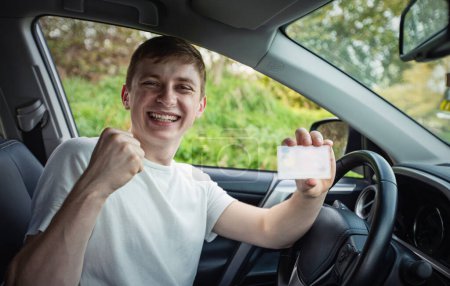 Un tipo feliz y orgulloso mostrando su licencia de conducir por la ventana del auto, mantiene apretado el puño como un ganador celebrando la victoria. Aprobación de la prueba y examen de conducir