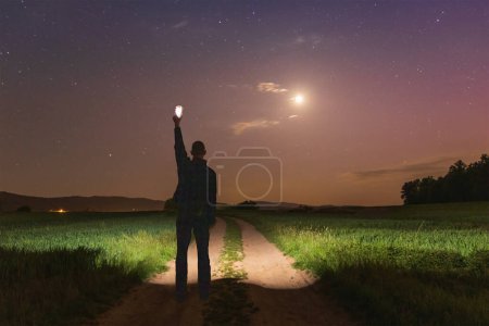 Man silhouette shining with mobile phone on way between field under night sky with star and moon