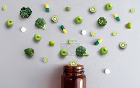 Foto de Pastillas y cápsulas de vitaminas verdes junto con manzanas, brócoli, col, lima y kiwi saliendo de un frasco de medicina, concepto de antioxidantes nutricionales - Imagen libre de derechos