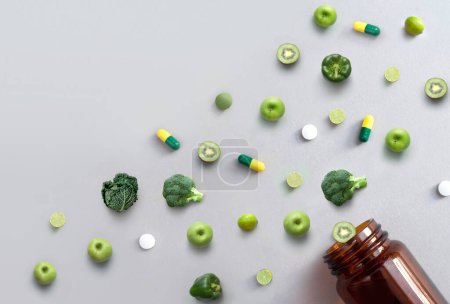 Photo for Green vitamin pills and capsules alongside apples, broccoli, cabbage, lime and kiwi coming out of a medicine jar, nutrition antioxidants concept - Royalty Free Image