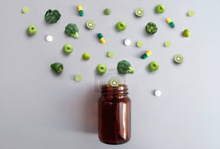 Téléchargez les photos : Pilules et capsules de vitamines vertes accompagnées de pommes, brocoli, chou, citron vert et kiwi sortant d'un pot de médicaments, concept d'antioxydants nutritionnels - en image libre de droit