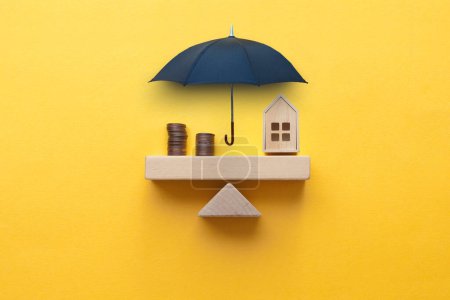 Photo for Umbrella protecting seesaw balance between house and coins, property insurance concept - Royalty Free Image