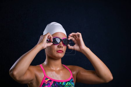 Photo for Pretty dark haired girl posing in a studio while wearing a training swimwear outfit that includes a swimsuit, swimming goggles, and a swimming cap. - Royalty Free Image