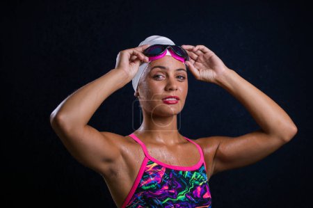 Foto de Chica de pelo bastante oscuro posando en un estudio mientras usa un traje de baño de entrenamiento que incluye un traje de baño, gafas de natación, y una gorra de natación. - Imagen libre de derechos