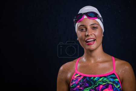 Photo for Pretty dark haired girl posing in a studio while wearing a training swimwear outfit that includes a swimsuit, swimming goggles, and a swimming cap. - Royalty Free Image