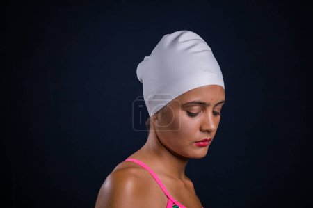 Foto de Chica de pelo bastante oscuro posando en un estudio mientras usa un traje de baño de entrenamiento que incluye un traje de baño, gafas de natación, y una gorra de natación. - Imagen libre de derechos