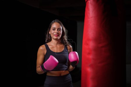 Photo for Pretty dark haired girl boxing a punching bag in a dark and moody setting. She is wearing boxing gloves and is fully focused on her training or exercise routine. - Royalty Free Image