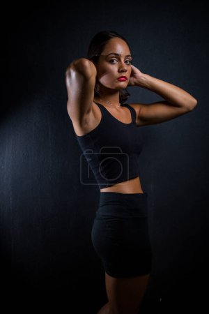 Foto de Chica de pelo bastante oscuro posando para una sesión de fotos de fitness en un estudio. Ella está vestida con pantalones cortos ajustados y una parte superior de la cosecha, y sus poses transmiten una sensación de fuerza, atletismo y aptitud. - Imagen libre de derechos