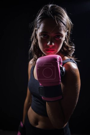 Foto de Chica bastante morena boxeando un saco de boxeo en un entorno oscuro y malhumorado. Lleva guantes de boxeo y está totalmente enfocada en su rutina de entrenamiento o ejercicio.. - Imagen libre de derechos