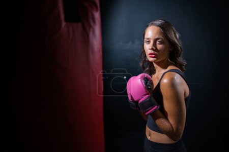 Téléchargez les photos : Jolie fille aux cheveux foncés boxant un sac de boxe dans un cadre sombre et lunatique. Elle porte des gants de boxe et se concentre entièrement sur sa routine d'entraînement ou d'exercice.. - en image libre de droit