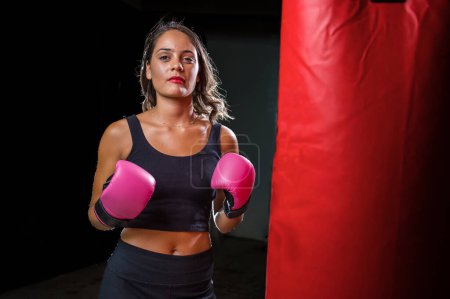 Photo for Pretty dark haired girl boxing a punching bag in a dark and moody setting. She is wearing boxing gloves and is fully focused on her training or exercise routine. - Royalty Free Image