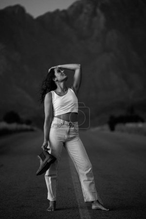 Téléchargez les photos : Jolie jeune femme aux cheveux bruns sur une route déserte. Elle est vêtue d'un vêtement tendance et à la mode, y compris un jean et un haut de culture élégant. - en image libre de droit