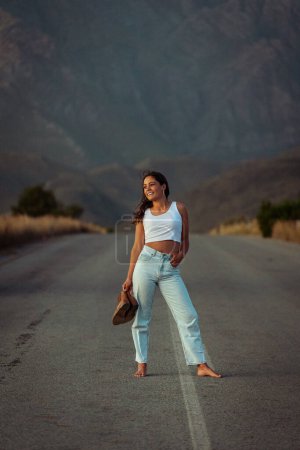 Photo for Pretty young woman with dark hair on a deserted road. She is dressed in trendy and fashionable attire, including jeans and a stylish crop top. - Royalty Free Image