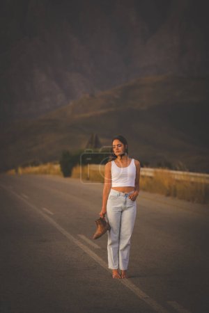 Téléchargez les photos : Jolie jeune femme aux cheveux bruns sur une route déserte. Elle est vêtue d'un vêtement tendance et à la mode, y compris un jean et un haut de culture élégant. - en image libre de droit