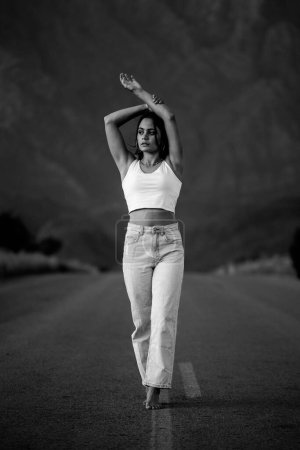 Photo for Pretty young woman with dark hair on a deserted road. She is dressed in trendy and fashionable attire, including jeans and a stylish crop top. - Royalty Free Image