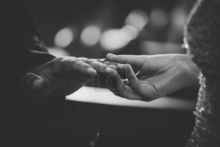 Dieses schöne Bild fängt den intimen Moment eines Paares ein, das bei einer echten Hochzeit Eheringe austauscht. Das Foto zeigt eine Nahaufnahme der Hände des Paares, die ihre ineinander verschlungenen Finger und die Hochzeitsbänder an ihren Fingern zeigt. Das Bild