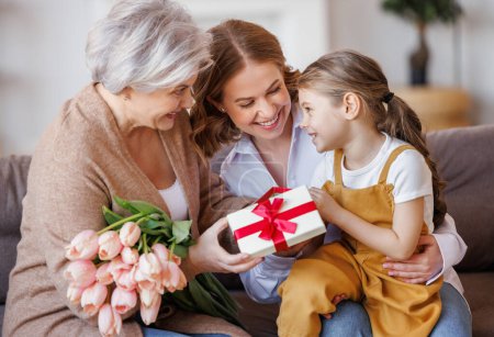 Photo for Happy International Women's Day. Smiling multi generational family daughter and granddaughter giving flowers  and gift to grandmother  cheerfully celebrate the spring holiday Mother's Day or grandparents day at home - Royalty Free Image