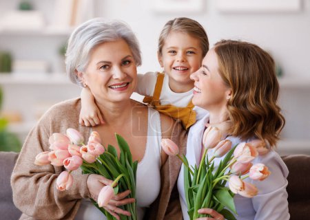 Téléchargez les photos : Joyeuse Journée internationale de la femme. Famille multi-générationnelle souriante grand-mère, mère et fille avec des fleurs fêtent joyeusement les vacances de printemps Fête des mères à la maison - en image libre de droit