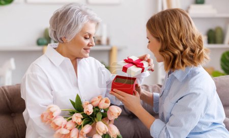 Photo for Positive elegant elderly woman with bunch of fresh tulips smiling and accepts a gift from a young daughter during a mothers day celebration - Royalty Free Image