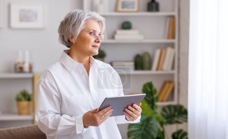 Foto de Mujer de mediana edad con tableta de navegación de pelo gris mientras mira enfocada en la pantalla en la sala de estar en casa - Imagen libre de derechos