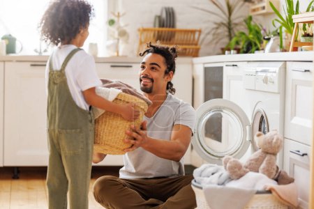 Téléchargez les photos : Garçon noir joyeux assis sur les genoux de papa et aidant père au linge dans le panier tout en faisant la lessive près de la machine à laver dans la cuisine de vol le week-end à la maison - en image libre de droit