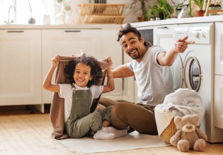 Téléchargez les photos : Optimiste père de famille noire et heureux fils en vêtements décontractés souriant et se couvrant de serviette tout en étant assis sur le sol et faire la lessive dans la cuisine à la maison - en image libre de droit