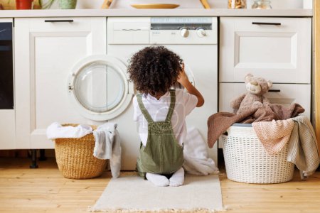 Foto de Vista posterior del chico negro con el pelo rizado arrodillado en el suelo y poner la ropa en la lavadora mientras se hace la colada en la cocina en casa - Imagen libre de derechos