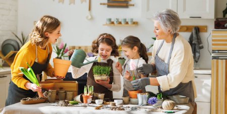 Téléchargez les photos : Grand-mère et mère positives replantant des fleurs de printemps dans des pots avec des enfants tout en se réunissant à table dans la cuisine domestique - en image libre de droit