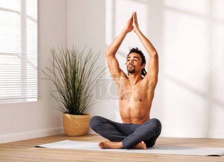 Photo for Calm African American man with naked torso sitting in Lotus pose with hands raised up  and meditating   in morning in sunlit room at home - Royalty Free Image